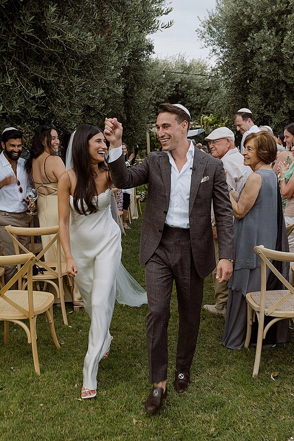 Wedding recessional as bride and groom walk the grass aisle, veil flowing, guests cheering under string lights in a garden setting