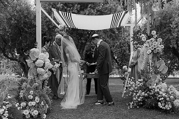 Wedding vows at an outdoor Jewish wedding ceremony as bride and groom hold hands under a striped chuppah with floral arch accents on a garden lawn