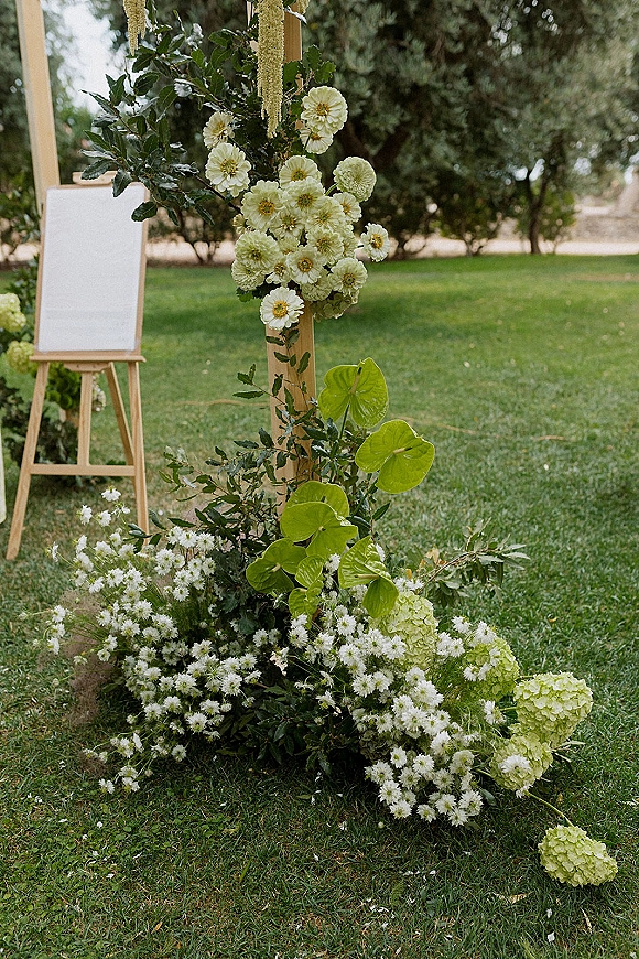 Wedding ceremony florals on a wooden wedding arch with white blooms, greenery and hanging tassels, set on a garden lawn with trees in back