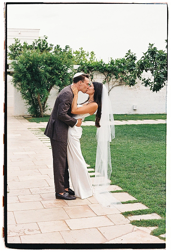 Wedding kiss as bride and groom kissing, her long veil flowing while he wears a dark suit and kippah in a garden by a white wall