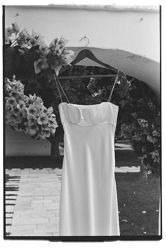Wedding dress with spaghetti straps on a hanger beneath an outdoor garden archway, framed by floral bushes and a stone walkway