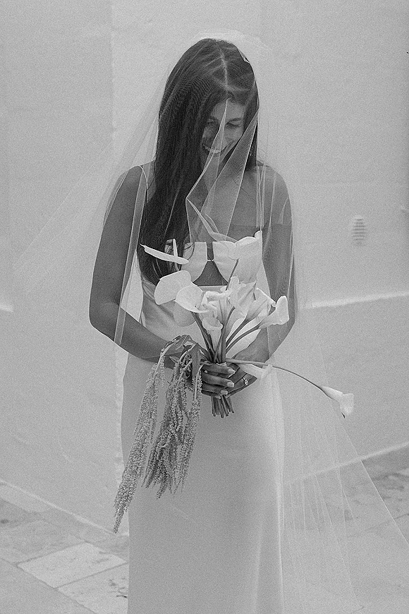Bridal portrait in black and white of a smiling bride with veil, looking down while holding a calla lily bouquet against a white wall