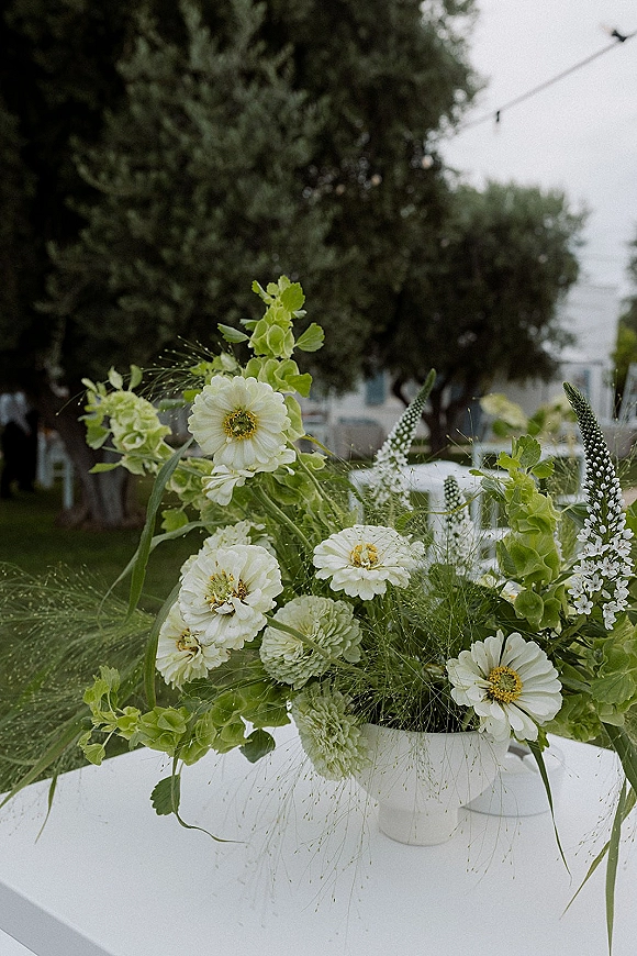 Wedding floral centerpiece of white flowers and greenery in a bud vase on a white tablecloth, with string lights over an outdoor lawn reception
