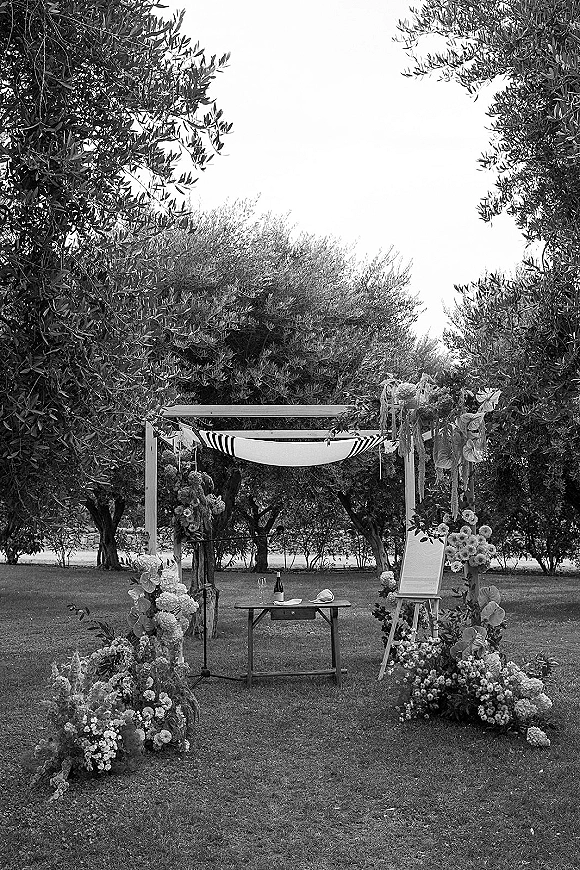 Ceremony setup with an outdoor wedding ceremony setup featuring a wood arch, fabric draping, lush florals, and olive trees on lawn grass