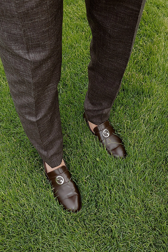 Groom shoes in brown leather loafers with monogram shoe clips beneath suit trousers, standing on a green grass lawn outdoors