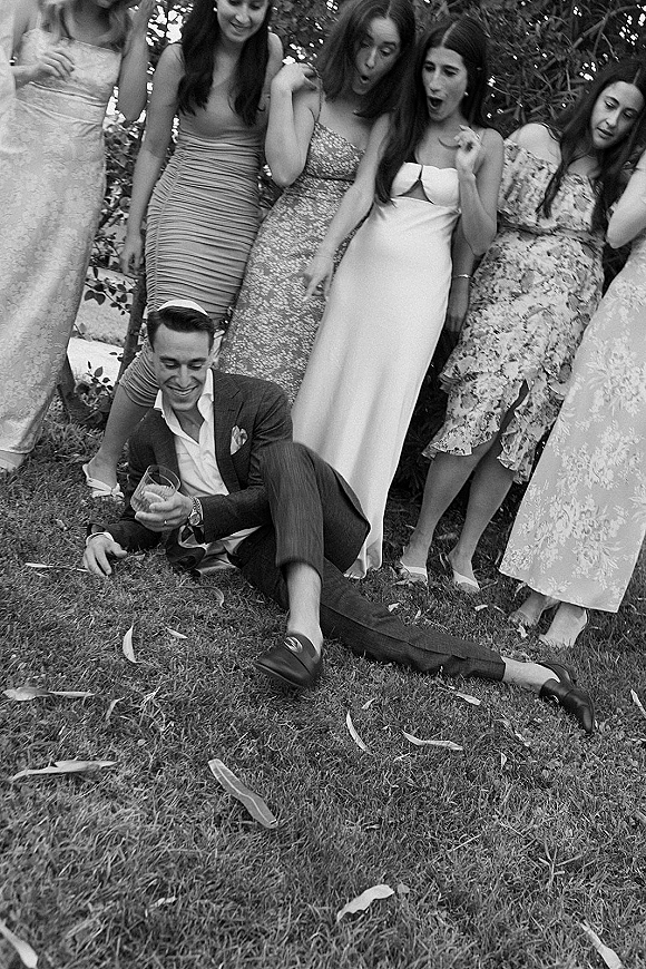 Wedding party photo of bridesmaids and groomsmen laughing on a grass lawn, groom seated with wine glass, floral dresses by stone steps