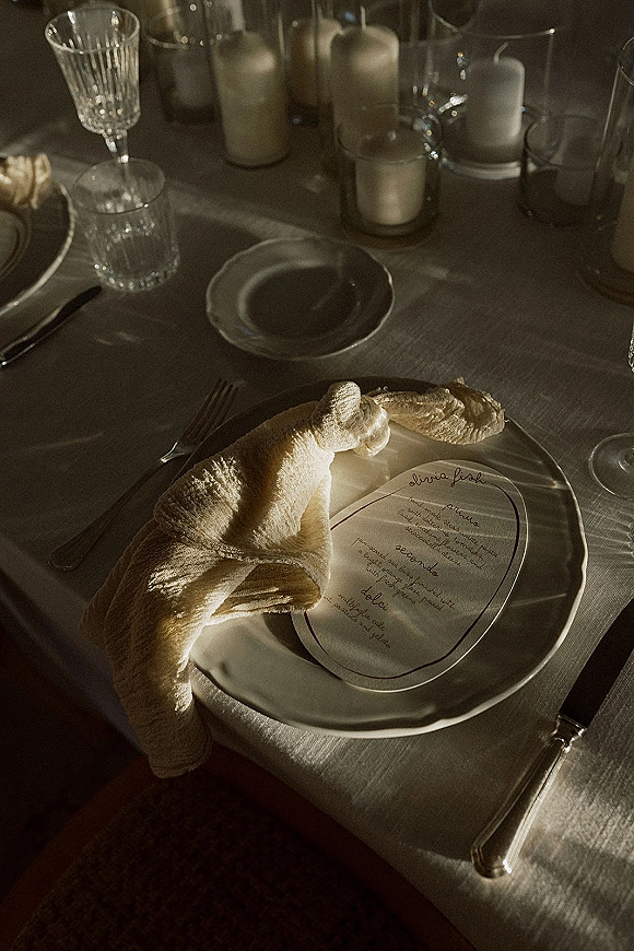 Wedding place setting with a linen napkin knot and round menu card on a white plate, silver flatware, champagne flute, and candlelight on table
