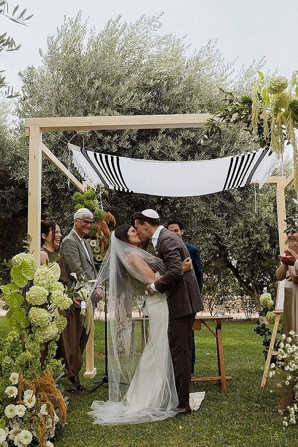 Wedding kiss under a chuppah canopy as guests applaud, bride in veil and gown with groom in suit amid hydrangea florals on a garden lawn