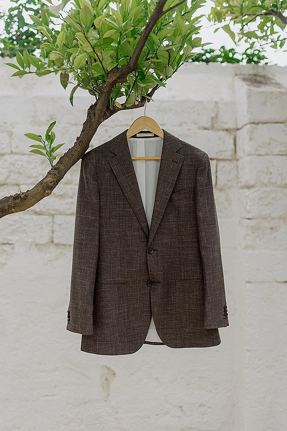 Groom suit hanging from a wooden hanger on a tree branch, suit jacket draped against a white stone wall with leaves nearby