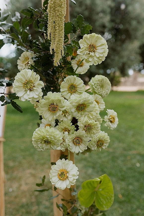 Wedding floral arrangement with white flowers and hanging tassel greenery on a wooden frame, set against a garden lawn and trees