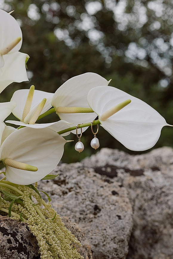 Wedding earrings with pearl drop earrings and gold hoops hanging on white anthurium flowers with greenery against stone rock and trees