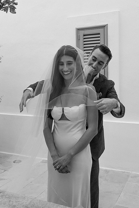 Wedding couple portrait with groom adjusting veil over smiling bride in strapless dress, posed by a white stucco wall and shuttered window