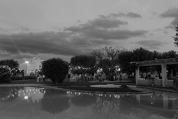 Outdoor wedding reception with backyard cocktail tables and lounge seating under string lights beside a pond and pergola on a cloudy evening