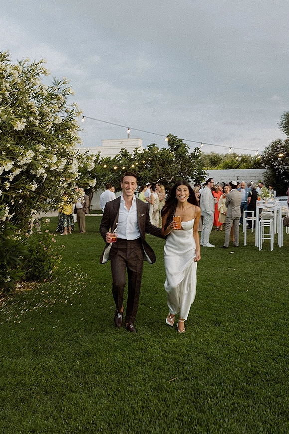 Reception couple moment as newlyweds walk in holding cocktails, bride in slip dress and groom in suit under string lights on a lawn at dusk