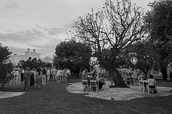 Outdoor wedding reception with wedding cocktail hour tables and chairs on a lawn under a large tree, accented by string lights and lanterns