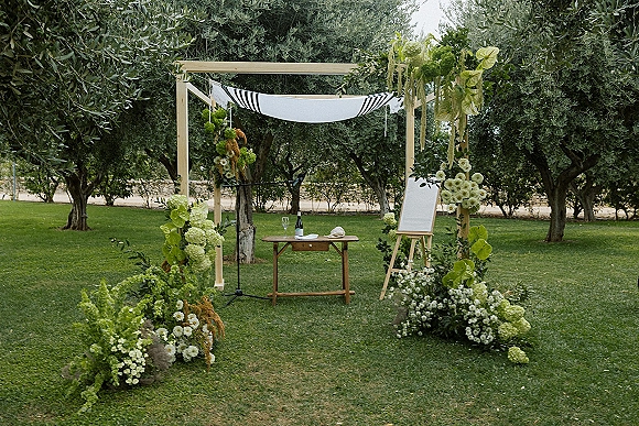 Ceremony altar decor with outdoor wedding altar featuring a wooden arch, green-white florals, draped striped canopy, and sign on easel on a garden lawn
