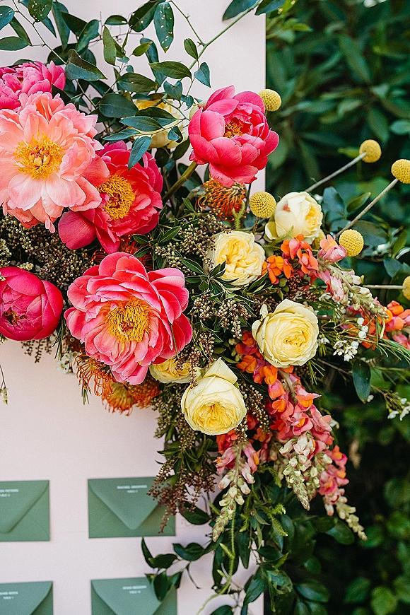 Wedding floral arrangement with peonies and roses, billy balls and greenery framing escort cards and envelopes on a display board, lush foliage behind