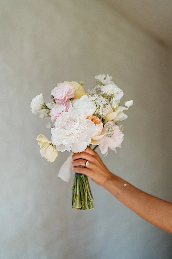 Bridal bouquet of white peonies and blush flowers, hand-tied with white ribbon, held by a bride showing an engagement ring against a neutral wall