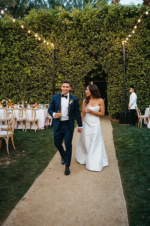 Newlywed couple walking hand in hand past outdoor reception tables, bride in strapless gown holding a cocktail, under string lights by a hedge wall