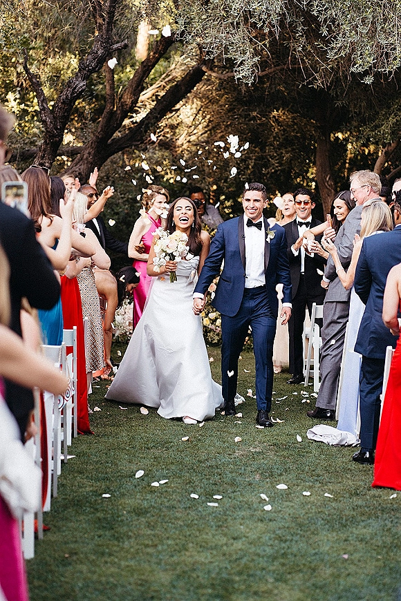 Wedding recessional as bride and groom walk the aisle holding hands while guests toss rose petals in an outdoor garden setting