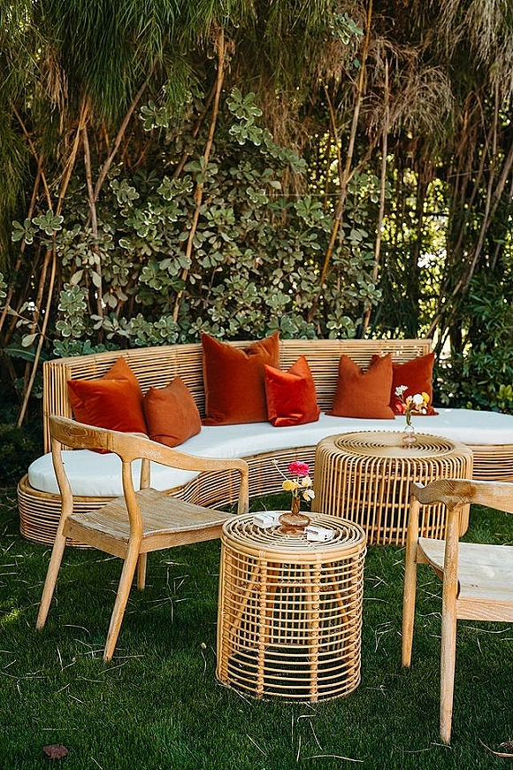 Outdoor lounge seating with rattan sofa, woven chair, and rust velvet pillows, styled with bud vases on a garden lawn under hanging foliage