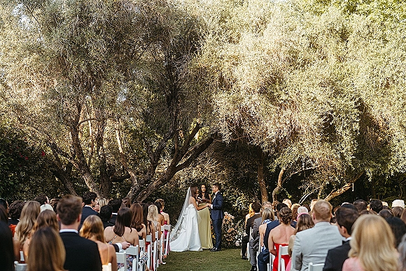Wedding vows as bride in veil and groom in suit stand with officiant by a floral altar on a garden lawn before seated guests