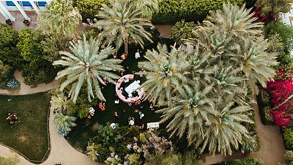 Outdoor wedding reception aerial view with round white linen tables, bar counter and cocktails, lounge chairs on a palm-lined garden lawn