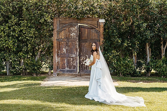 Bridal portrait of a bride holding bouquet in a strapless wedding dress with cathedral veil, smiling by wooden doors and garden trees