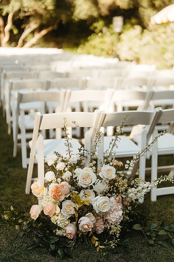 Ceremony aisle decor with outdoor ceremony seating, white folding chairs and a rose-hydrangea floral arrangement on a garden lawn by a tent