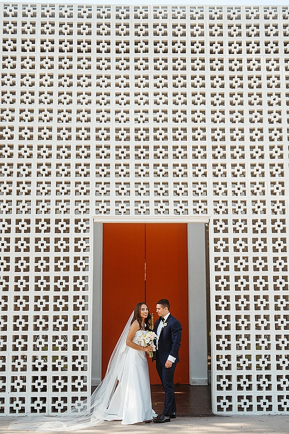 Couple portrait of bride and groom portrait with her bouquet and cathedral veil, posed by red double doors and patterned concrete wall