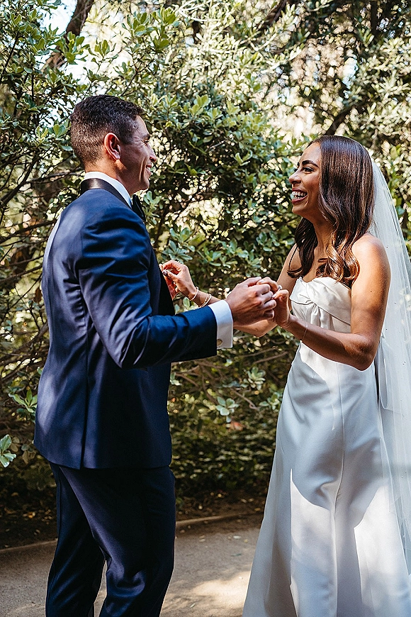 First look moment with bride and groom holding hands, her long veil trailing as they laugh on a sunlit garden path with trees