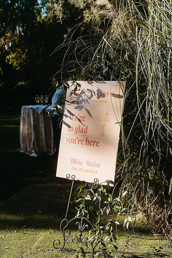 Wedding welcome sign with names in modern calligraphy and a cocktail illustration on a metal stand with pampas grass on a garden lawn