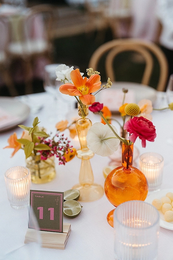 Reception tablescape with wedding table centerpiece of bud vases and colorful flowers, amber glass vase, citrus slices, and candlelight on white linen table