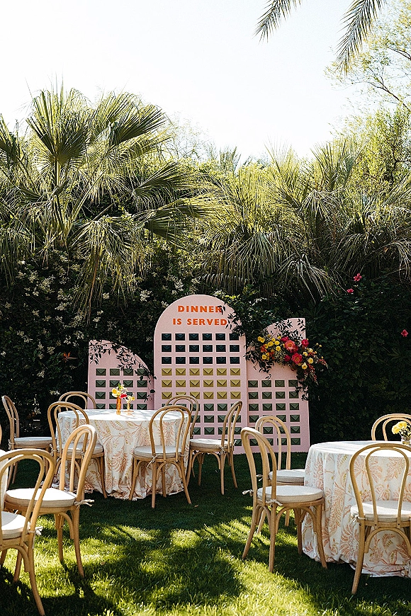 Wedding seating chart with escort card display and envelope cards on a lawn, framed by palm trees and garden reception tables with florals