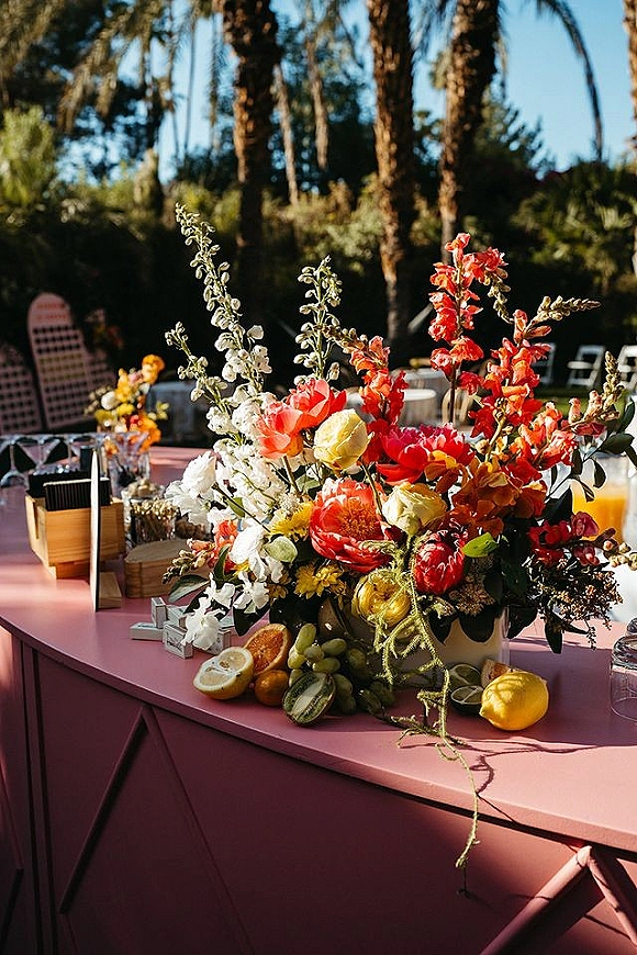 Reception tablescape with a wedding table centerpiece of colorful florals and citrus on a pink tablecloth, with candles in a sunlit garden setting