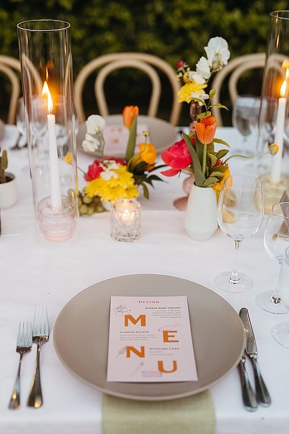 Reception tablescape with wedding place setting, menu card on charger plate, wine glasses and taper candles beside colorful flowers and greenery