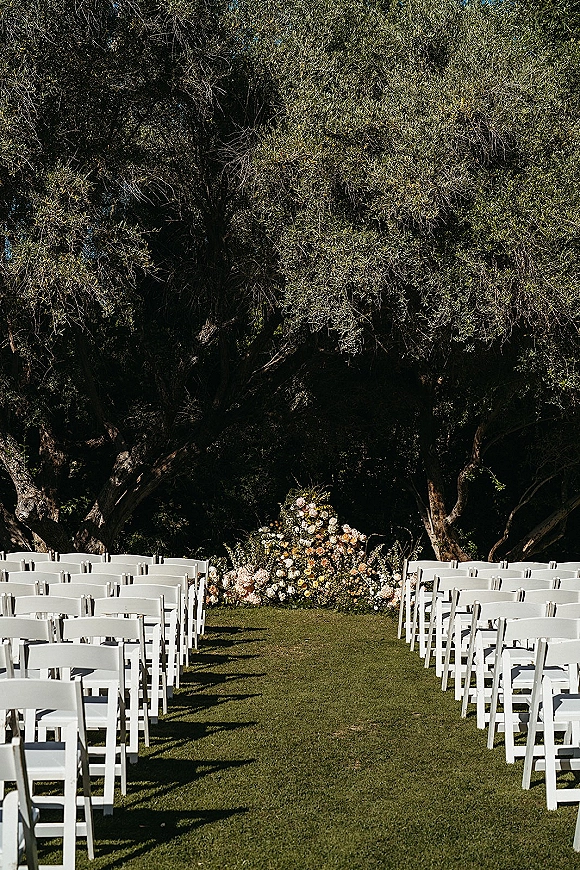 Ceremony setup for an outdoor wedding ceremony with rows of white folding chairs on a lawn, centered on a low rose floral arrangement under trees
