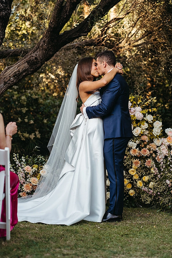 Wedding kiss portrait of bride and groom kiss in a garden, her strapless satin dress and cathedral veil beside a rose arrangement