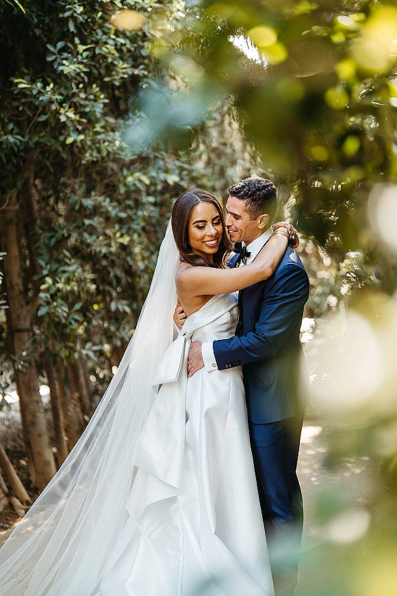 Wedding couple portrait of bride and groom embrace in a lush garden, her long veil draping as he kisses her forehead in a navy suit