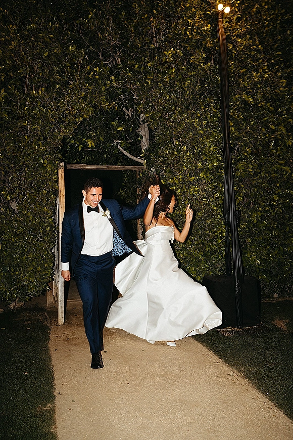Newlywed exit as bride and groom walk hand in hand, her strapless ball gown twirling beside his navy tuxedo by a garden gate and hedge wall