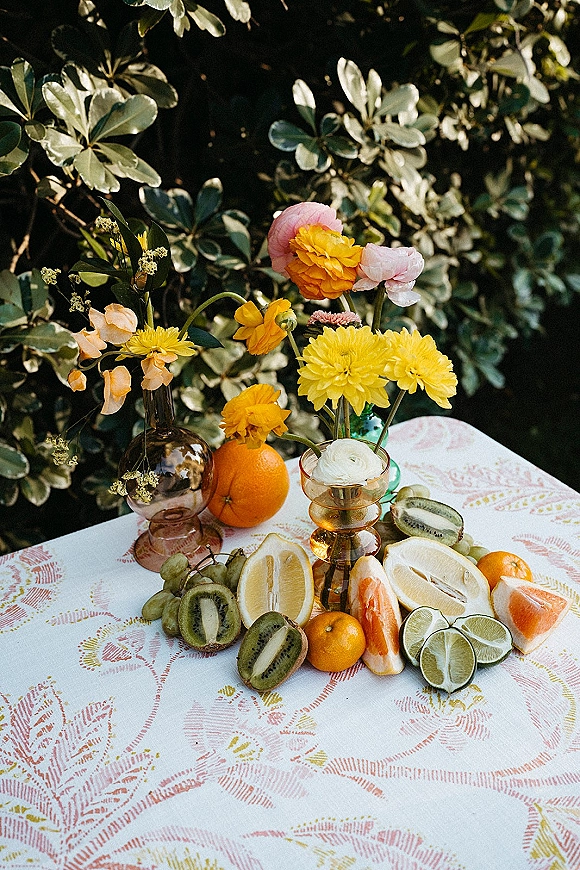 Reception tablescape with citrus wedding centerpiece, amber bud vases, yellow and pink blooms, and fruit on a patterned tablecloth by garden greenery