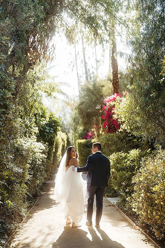 Couple portrait of bride and groom walking away holding hands, bride looking back as veil glows on a sunlit garden path with bougainvillea