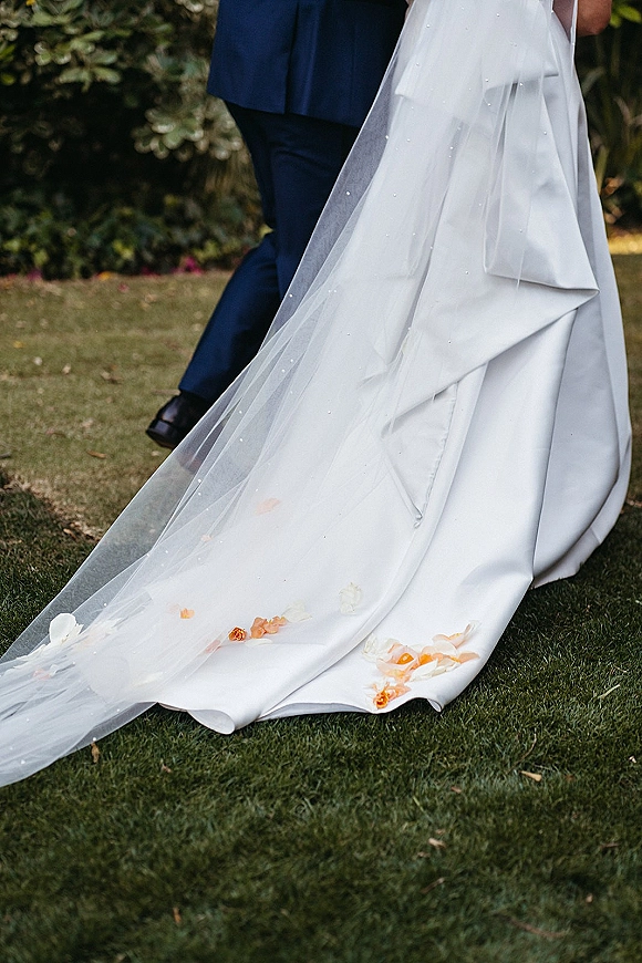 Wedding dress train with a cathedral veil train trailing over flower petals as the couple walks on a grassy garden lawn