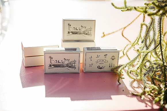 Wedding matchboxes with custom wedding matchboxes monogram label and venue illustration, styled with greenery on a pink tabletop in sunlight