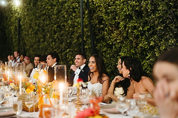 Reception moment at a wedding reception dinner as the bride and groom laugh with bridesmaids at a long banquet table before a hedge wall
