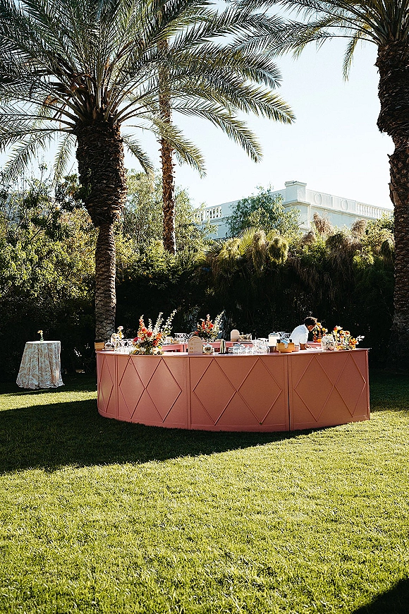 Outdoor wedding bar with a pink accent, floral arrangements, glassware and bottles on a curved setup beneath palm trees on the lawn