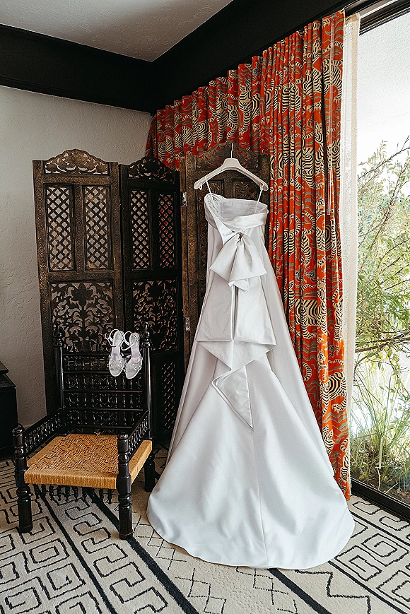 Wedding dress hanging in window light, satin wedding dress draped on a hanger beside bridal shoes near a carved folding screen and curtains
