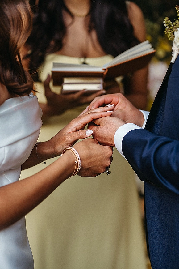 Ring exchange as the bride places a wedding ring exchange on the groom’s finger, close-up hands with bracelets and officiant book outdoors