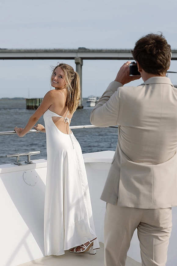 Bridal portrait of a bride on boat, looking back in an open-back white dress as the groom photographs her on a deck by water and bridge