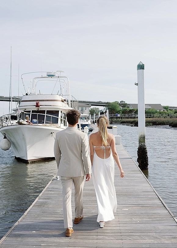 Couple portrait of bride and groom walking away on a dock at a marina, her open-back wedding dress and his suit by the water and yacht